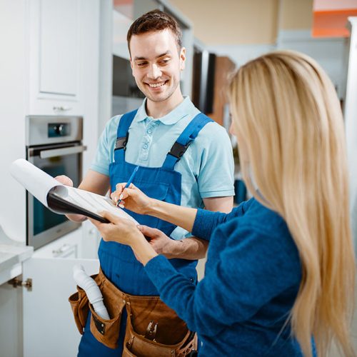 Male plumber in uniform and female customer in the kitchen. Handyman with toolbag repair sink, sanitary equipment service at home