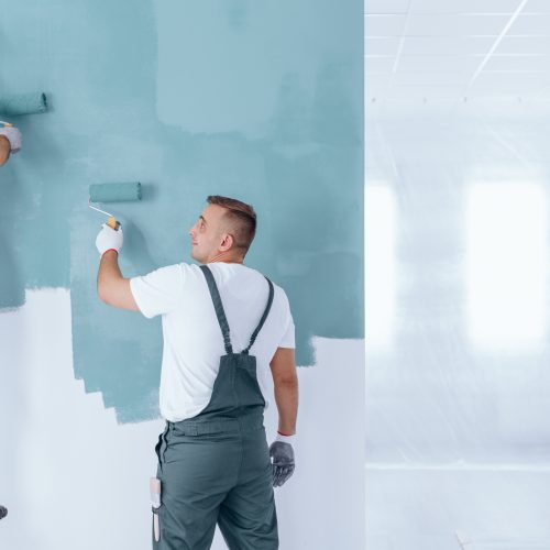 Men painting the wall blue using rollers in empty home interior